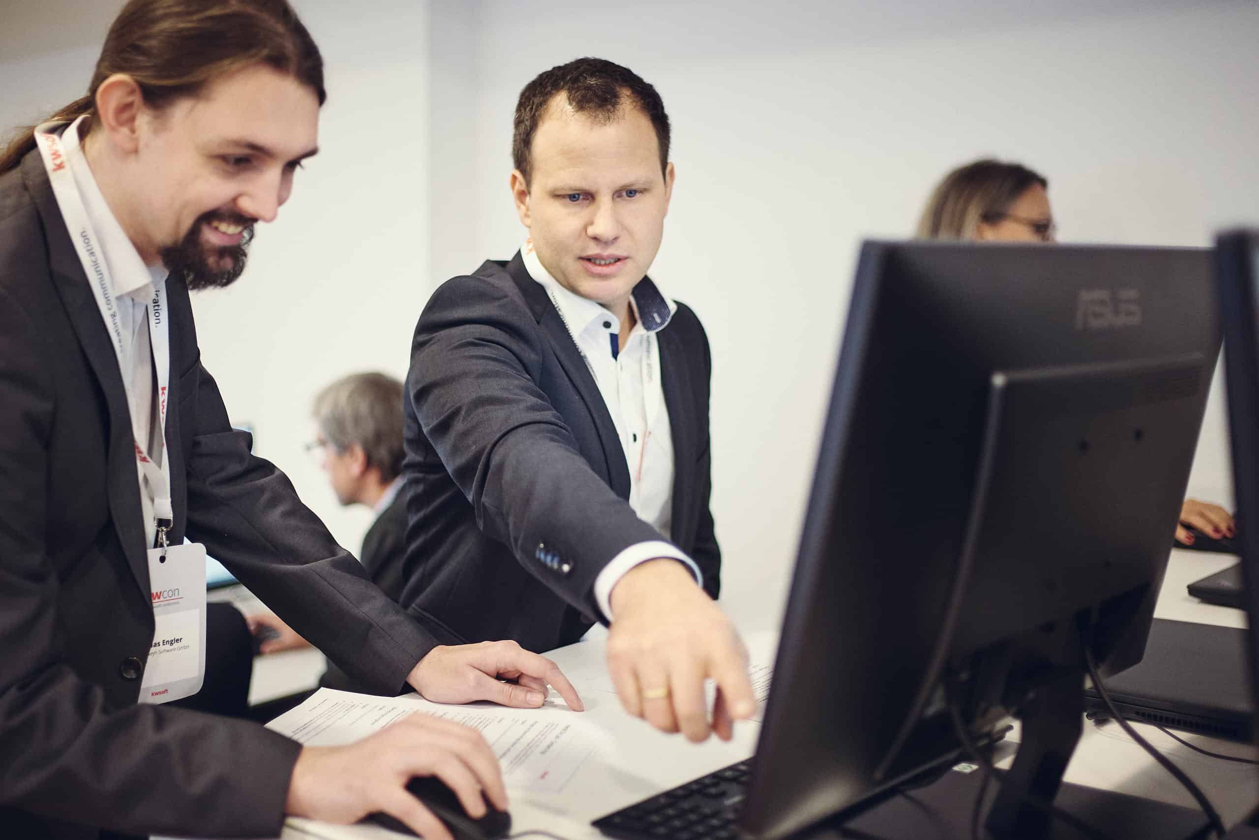 Dwóch mężczyzn w garniturach patrzy na ekran; ten po prawej wskazuje na ekran, a drugi obsługuje myszkę.